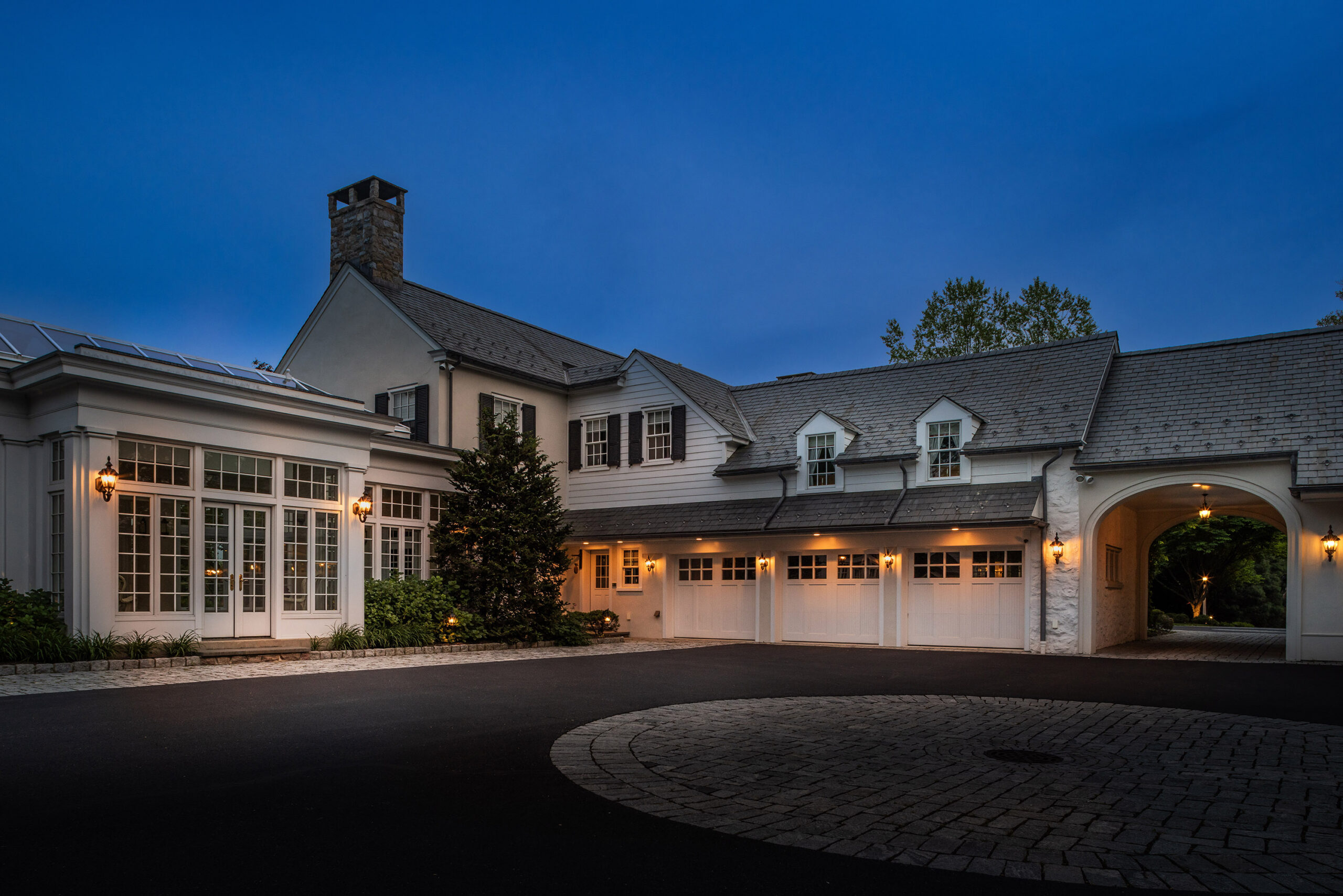 The forecourt of a grand white home at dusk. The home has many outdoor lights turned on.